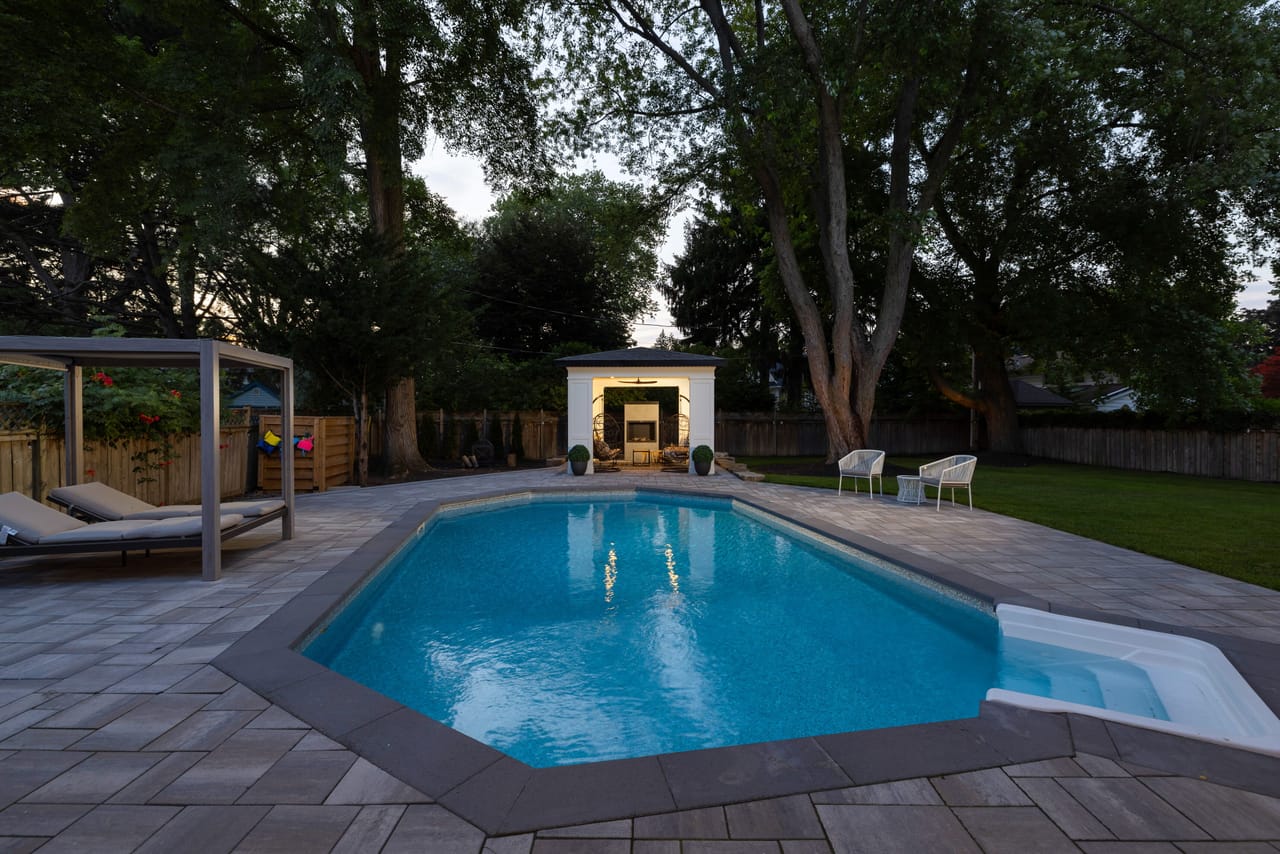 Backyard pool at twilight beside the cabana, with loungers under a pergola and an outdoor fireplace