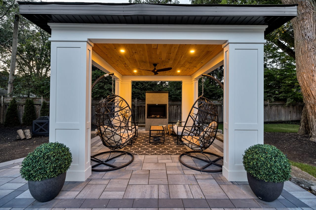 Covered cabana at dusk, hanging woven chairs and a gas fireplace under a cedar-clad ceiling