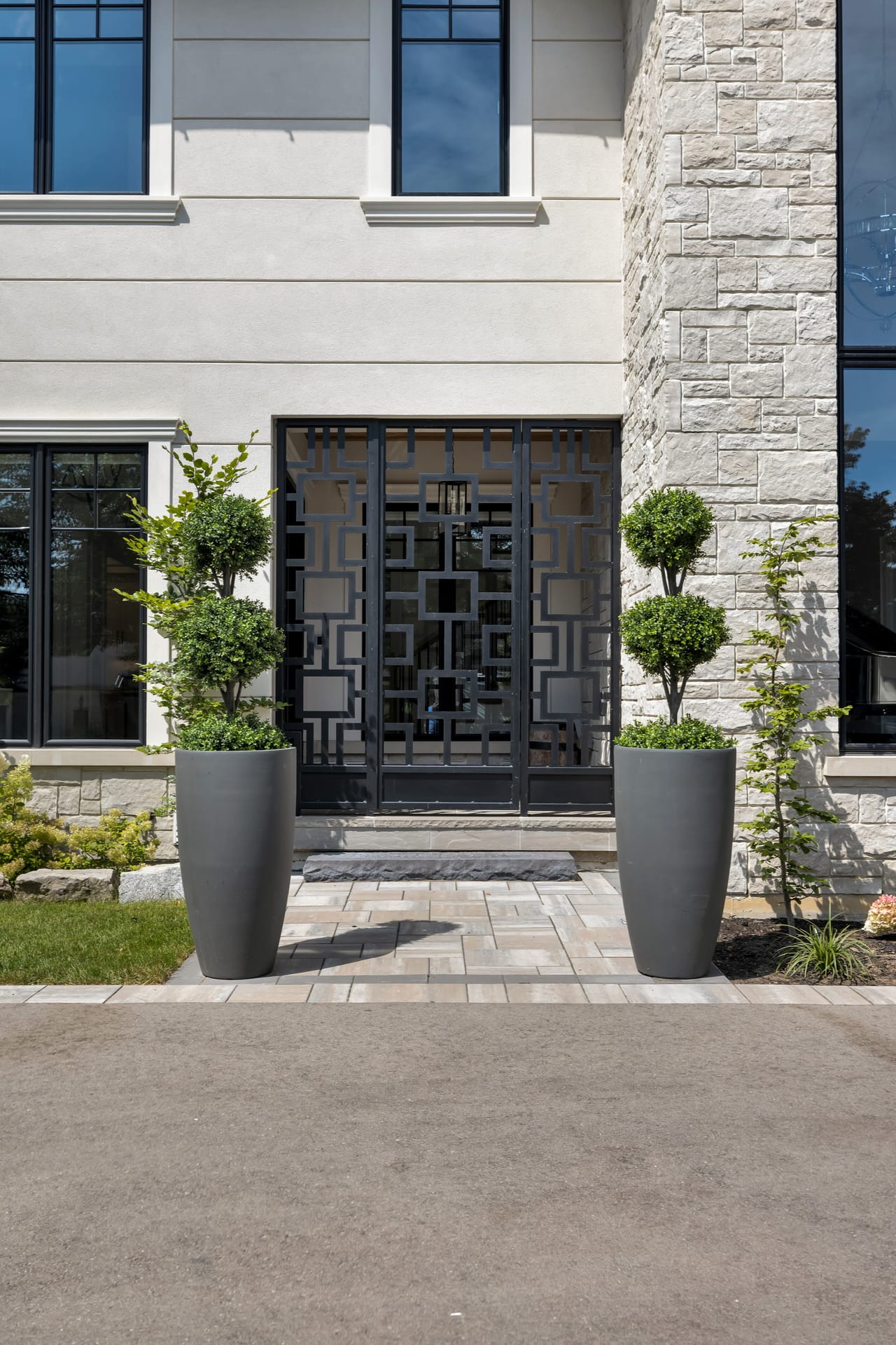 Geometric metal entry doors flanked by tall topiaries in charcoal planters