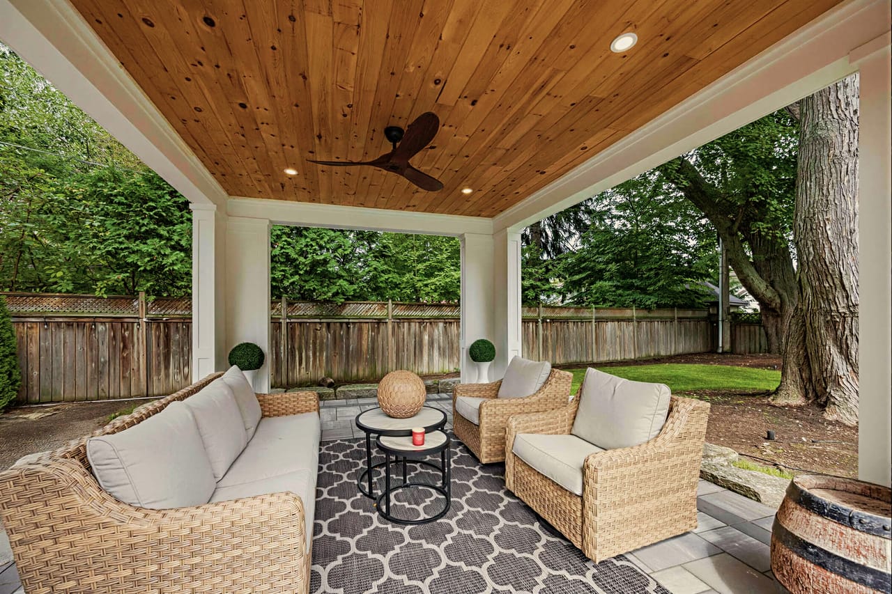Covered loggia under a cedar plank ceiling, deep wicker seating facing the lawn
